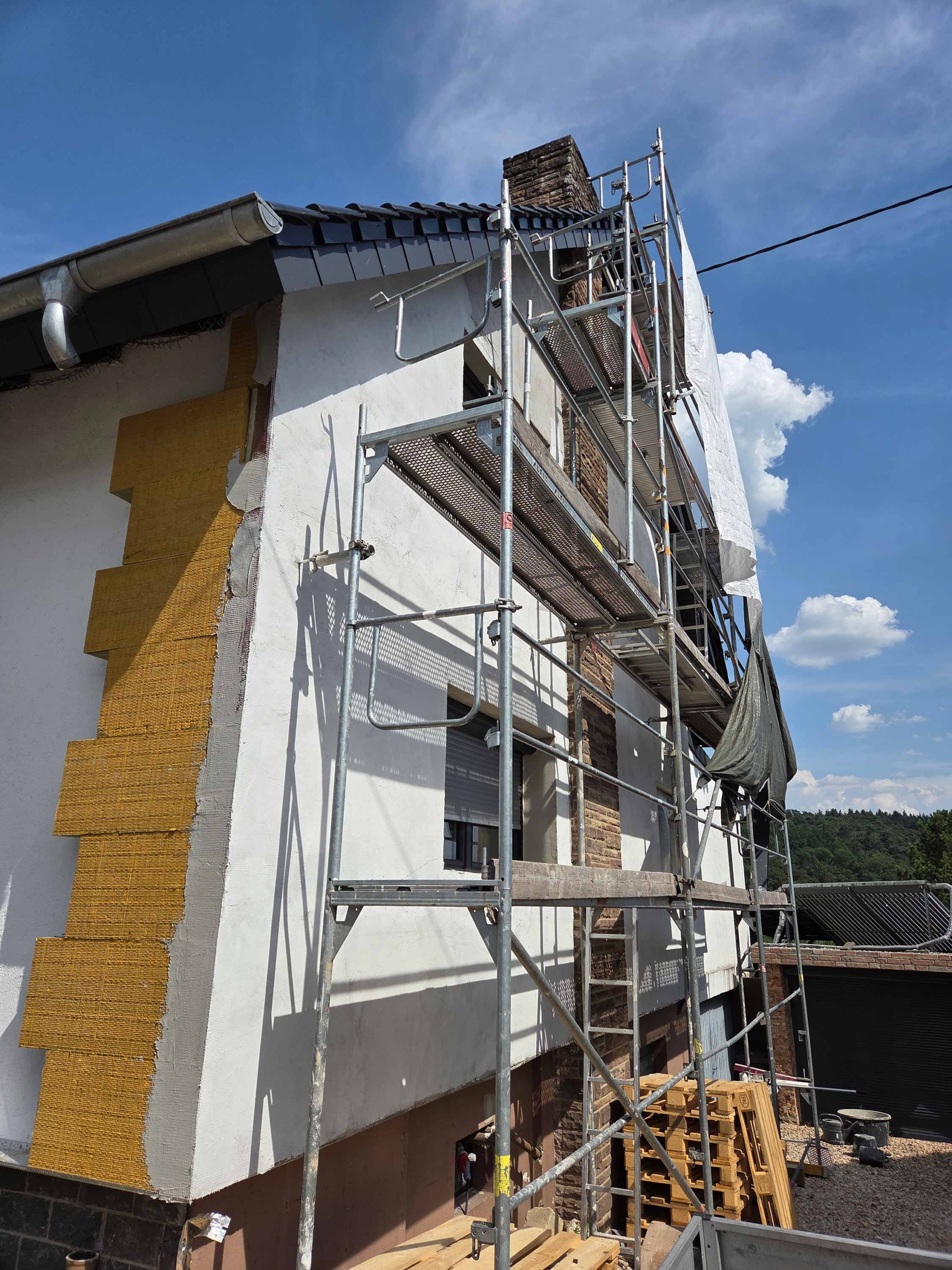 Ecke der Fassade eines Einfamilienhauses. Bei der linken Seite sieht man eine Reihe Dämmplatten, die rechte Seite ist bereits fertig verputzt. Dort steht noch ein Gerüst.